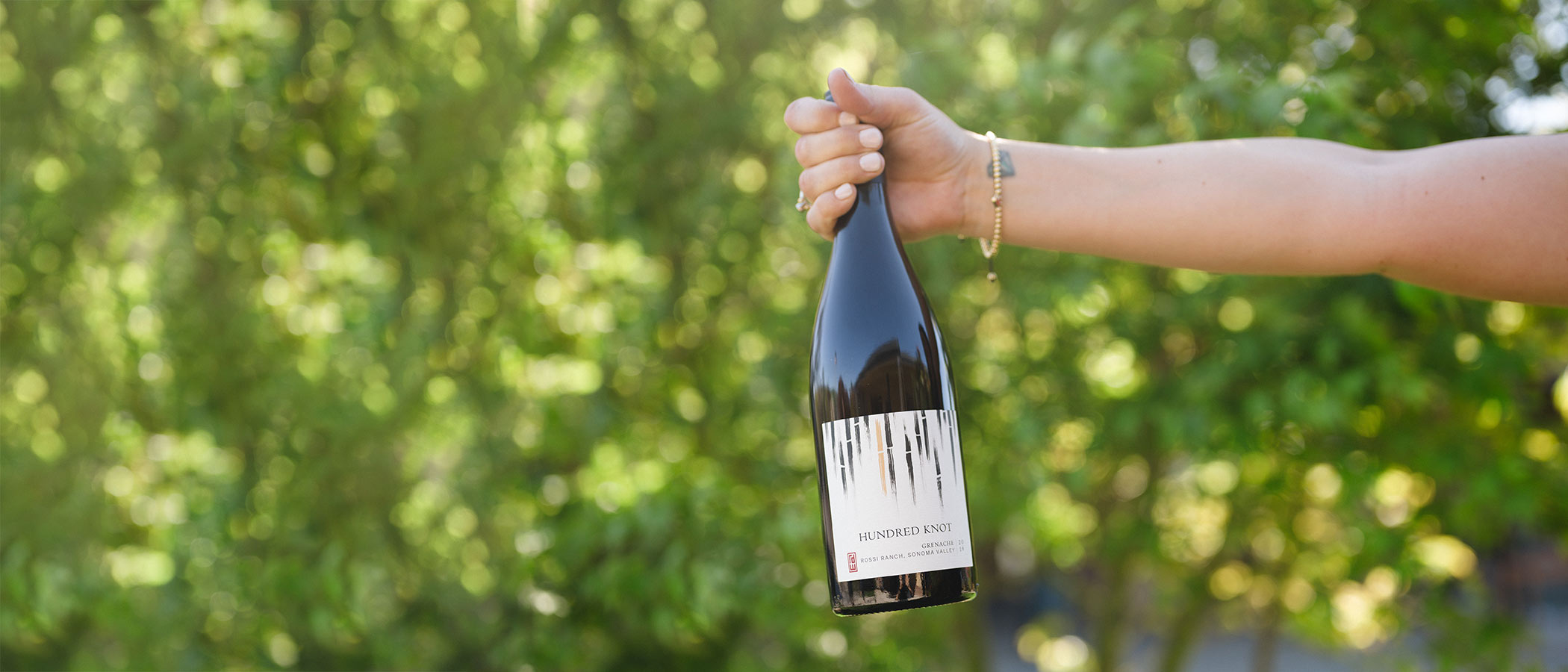 Hand holding RD Winery Hundred Knot Grenache in front of bushes.