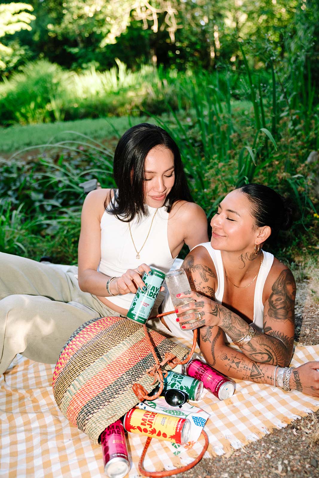 two women on a picnic blanket toasting with a glass of sparkling water and a can of wine spritzer with a basket turned on its side with cans spilling out