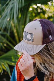 A woman outdoors wearing a beige and dark maroon trucker hat with a rectangular patch on the front. The hat has a beige front panel and brim, dark maroon mesh back panels, and a small white logo on the side.
