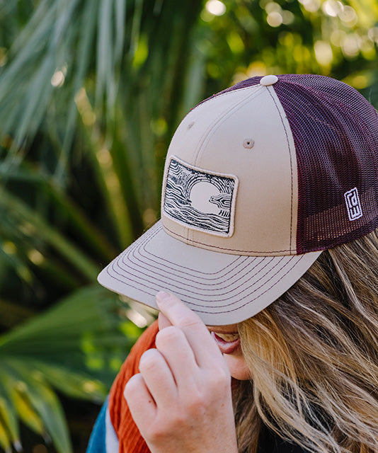 A woman outdoors wearing a beige and dark maroon trucker hat with a rectangular patch on the front. The hat has a beige front panel and brim, dark maroon mesh back panels, and a small white logo on the side.
