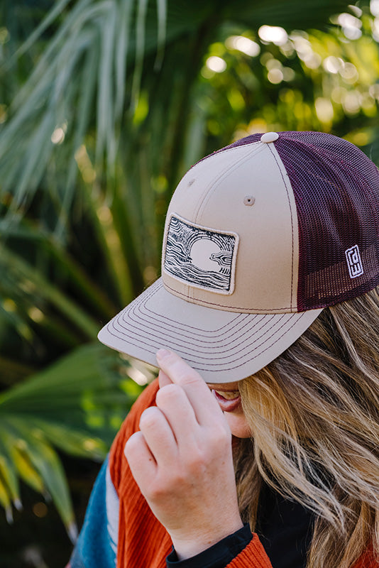 A woman outdoors wearing a beige and dark maroon trucker hat with a rectangular patch on the front. The hat has a beige front panel and brim, dark maroon mesh back panels, and a small white logo on the side.