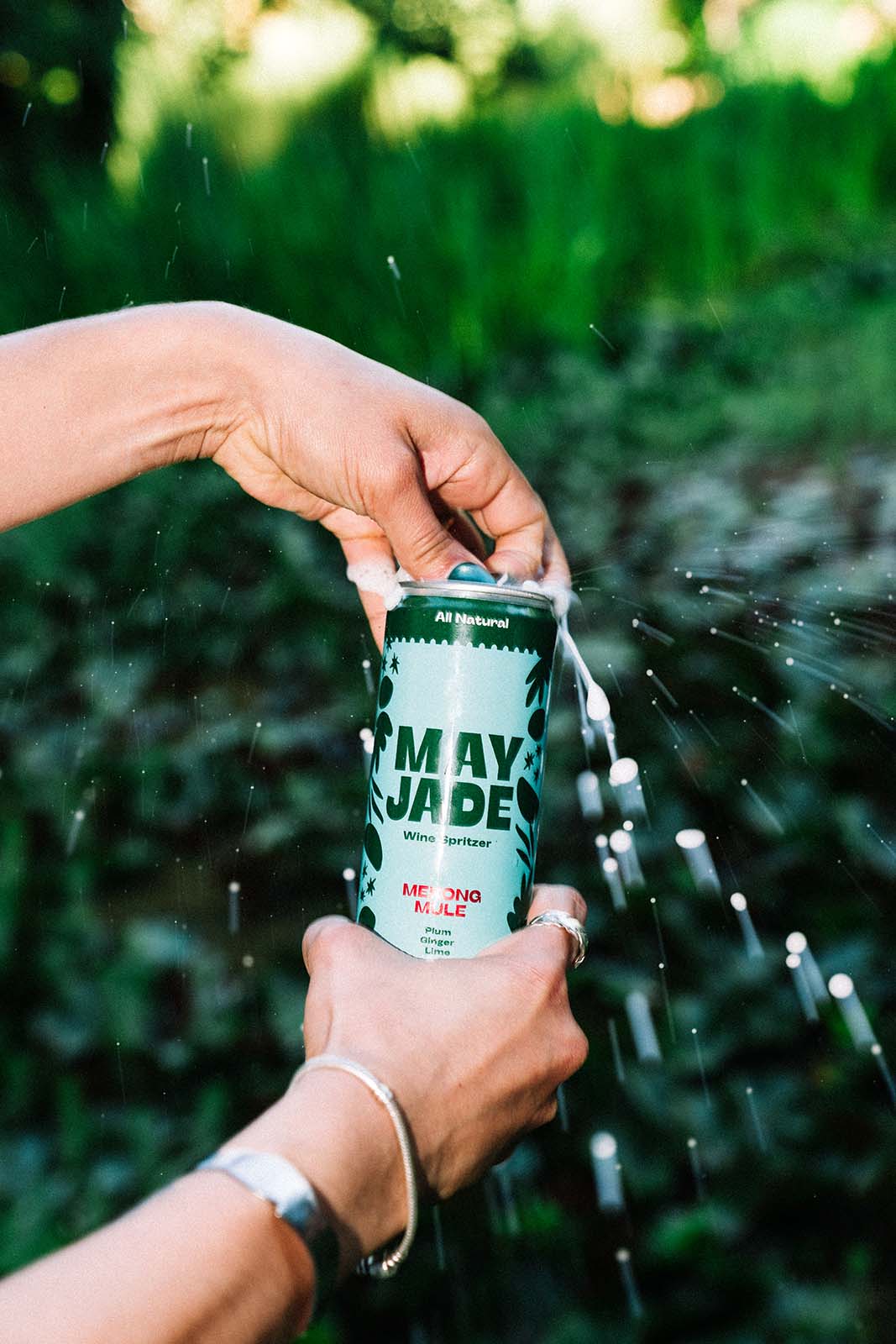 A person's hands holding a can of May Jade wine spritzer against a blurred green outdoor background with water spraying around the can.