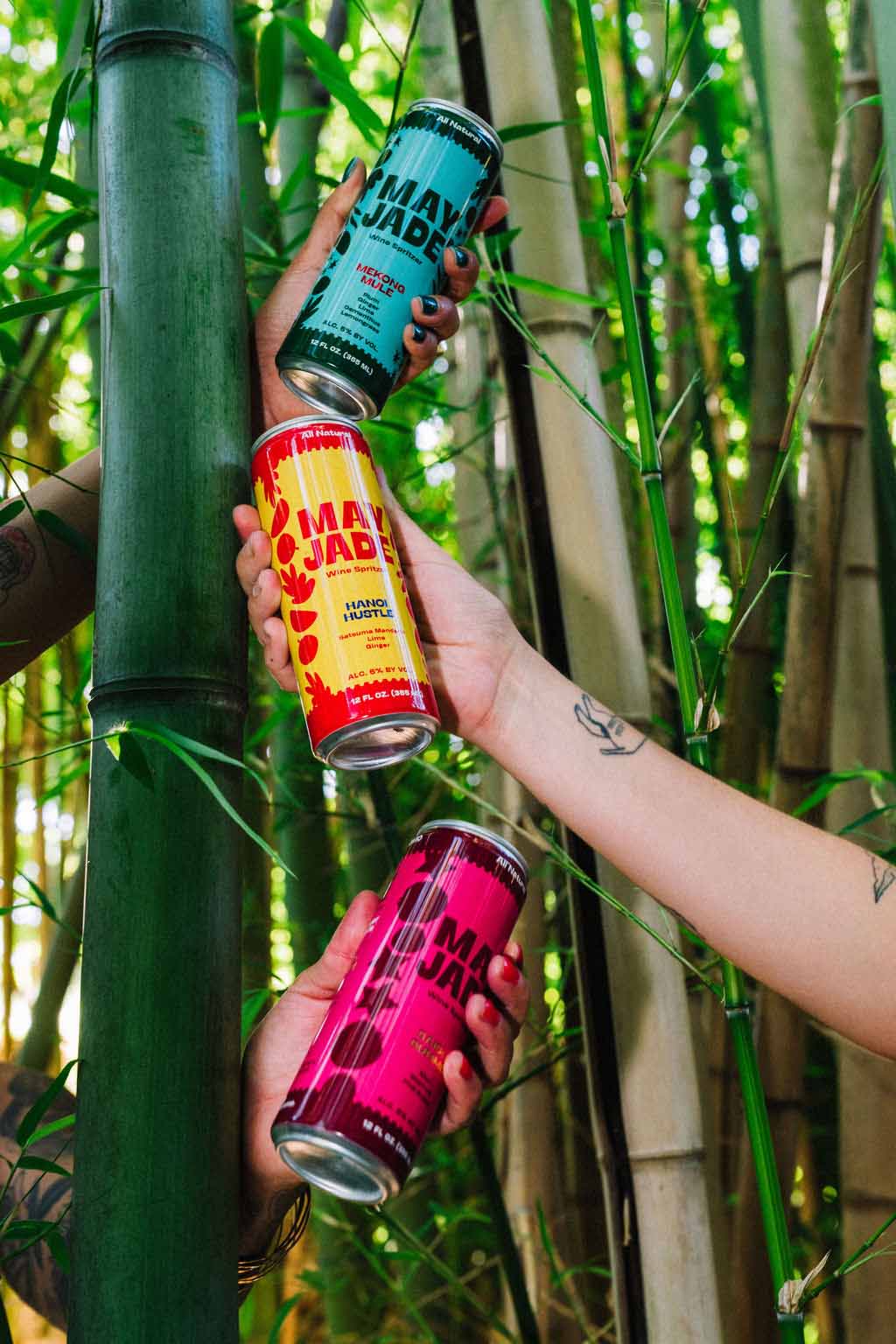 Three hands holding colorful wine spritzer cans in front of a bamboo forest. The cans are branded May Jade, with one can in teal, one in yellow and red, and one in pink.