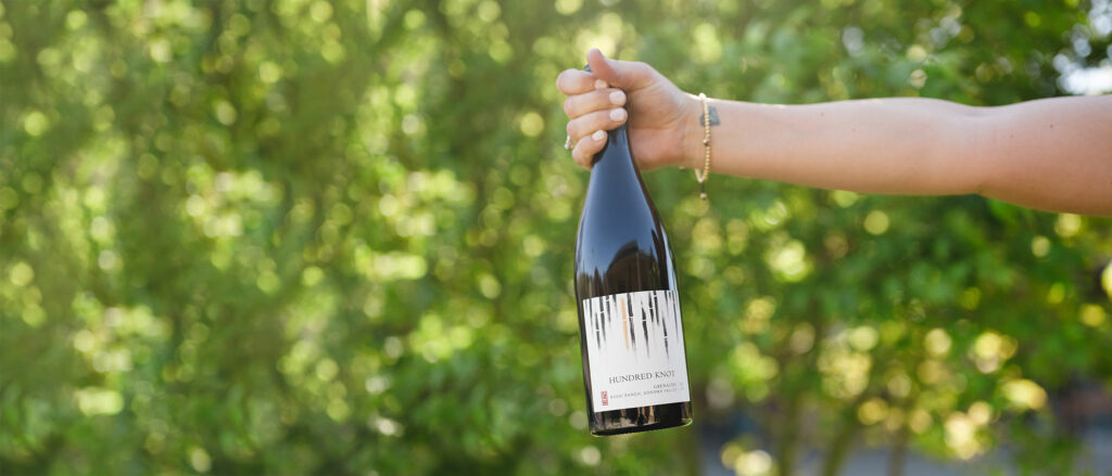 Hand holding RD Winery Hundred Knot Grenache in front of bushes.
