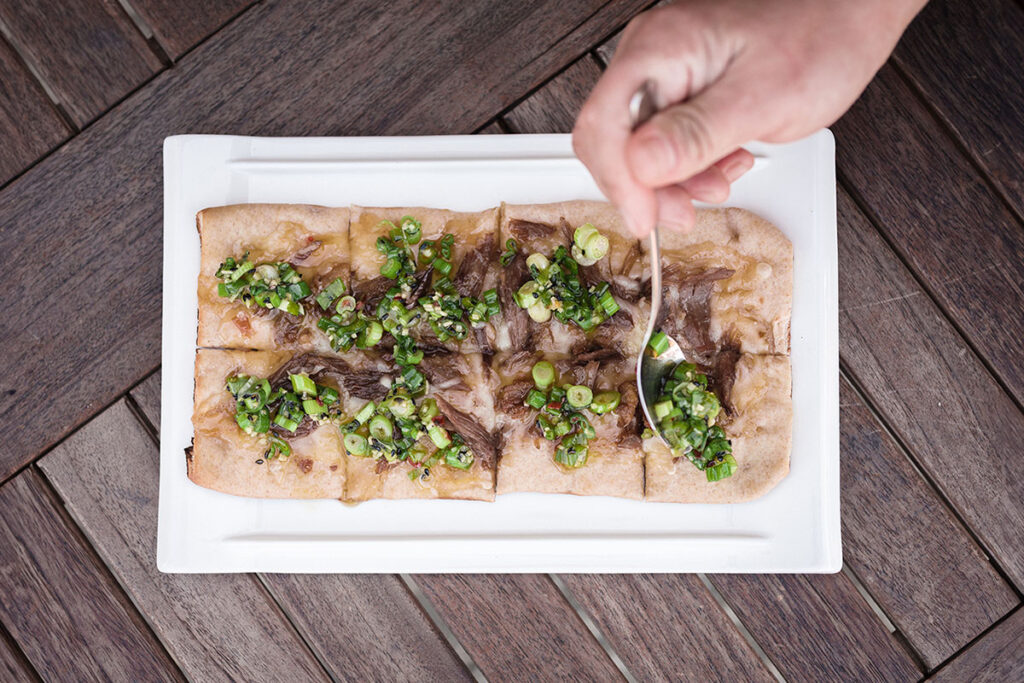 Overhead shot of person garnishing duck confit flatbread on wooden table