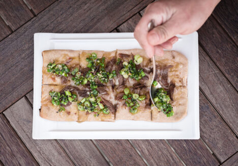 Overhead shot of person garnishing duck confit flatbread on wooden table