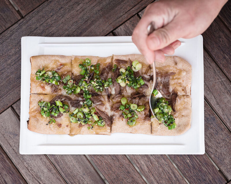 Overhead shot of person garnishing duck confit flatbread on wooden table